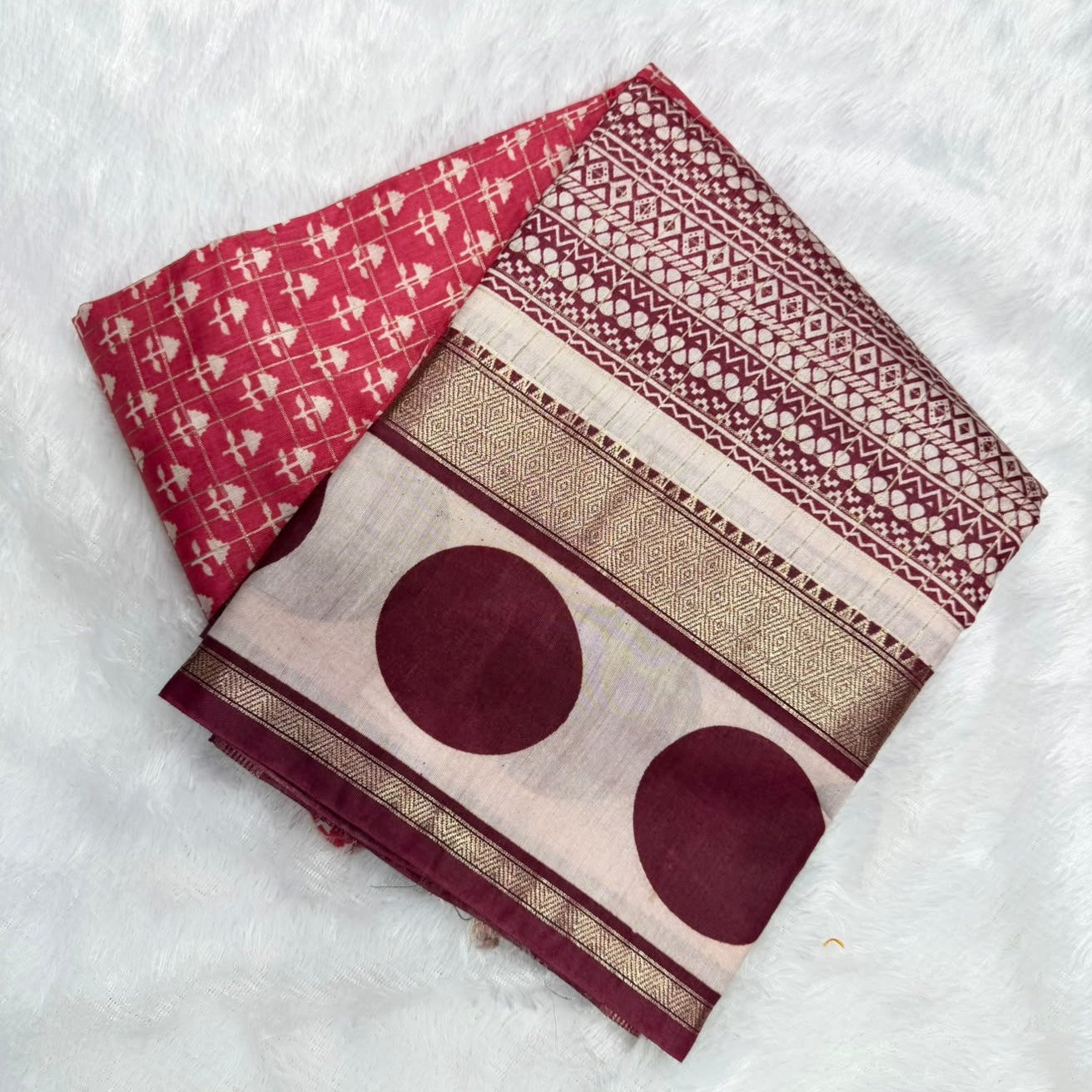 A folded traditional Indian saree displayed on a white faux fur background. The saree features a deep red body with white geometric patterns, a beige and maroon border with gold zari detailing, and two large maroon circles printed on the reverse side.