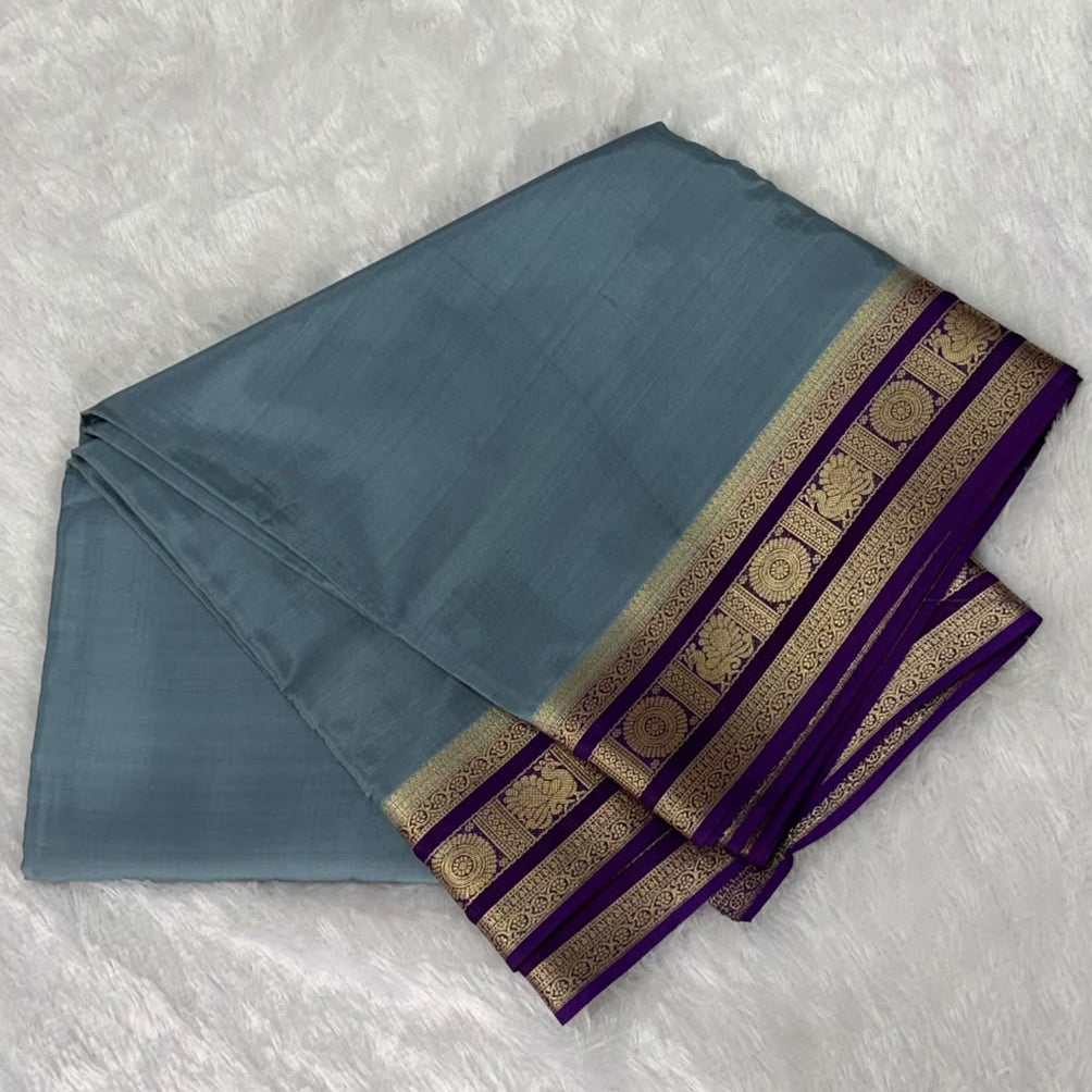 A folded slate blue saree or fabric piece with a purple and gold patterned border, laid flat on a white textured background.