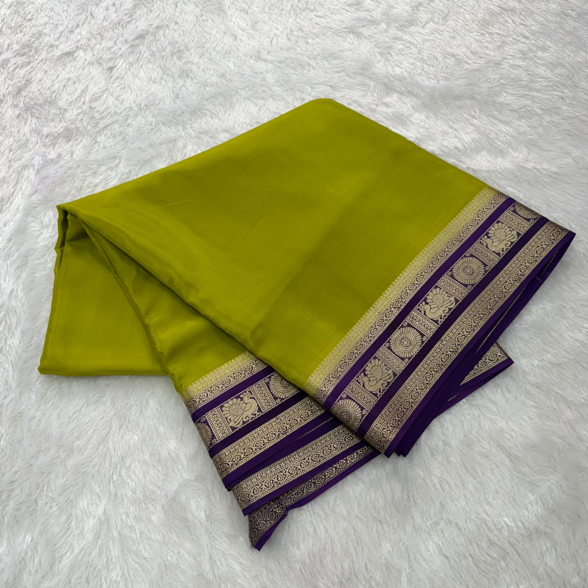A folded olive green saree or fabric piece with a purple and gold patterned border, displayed on a white faux fur background.