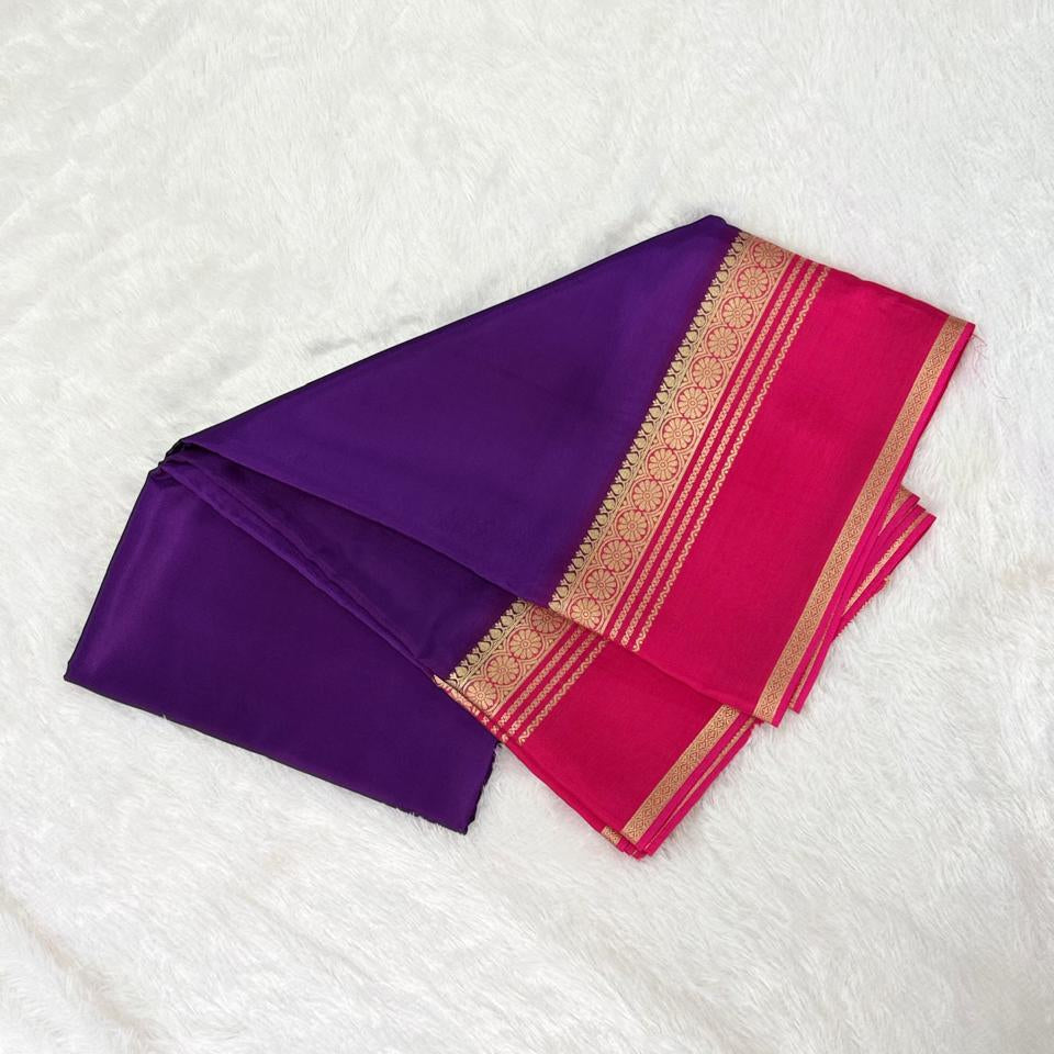 A folded purple and magenta saree with a gold woven border, displayed on a white faux fur background.