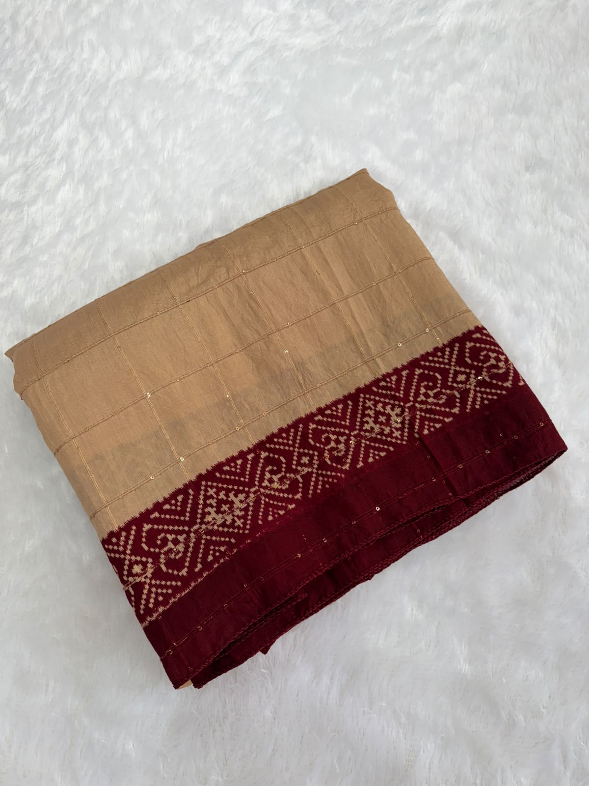 A folded beige and deep red saree or fabric piece with a detailed geometric border pattern, laid flat on a white textured faux fur background.