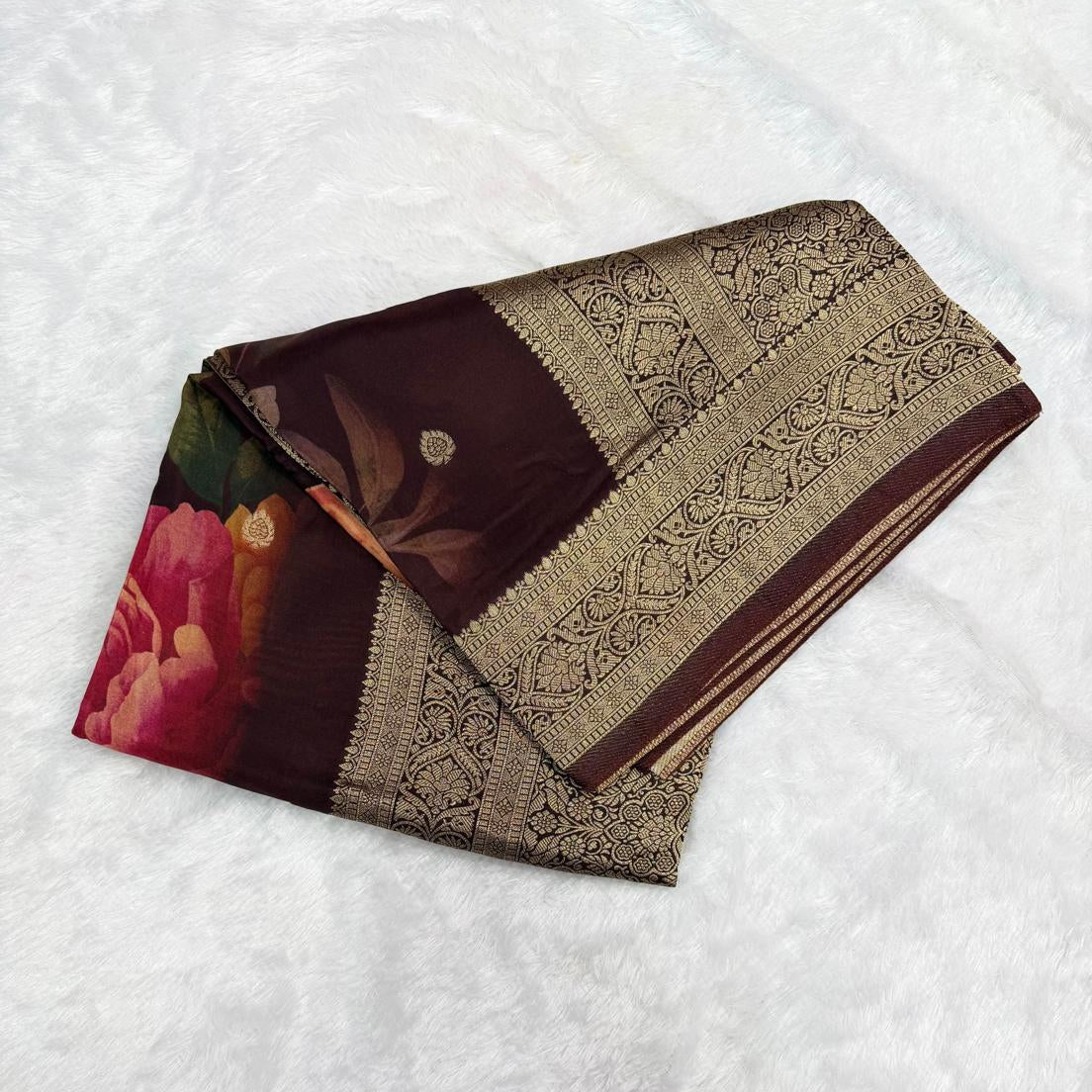 A folded dark brown silk saree with a wide beige and gold brocade border and a pink floral print on the end, displayed on a white textured fabric background.